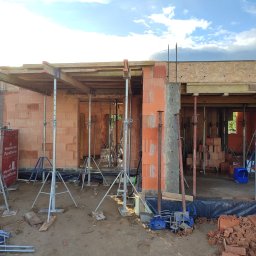 AWP-Domy Justyna Osowiecka - Budowa domu z czerwonej cegły Porotherm w Warszawie, widoczne ściany w trakcie murowania, drewniane szalunki podtrzymywane przez metalowe podpory, na ziemi porozrzucane cegły.