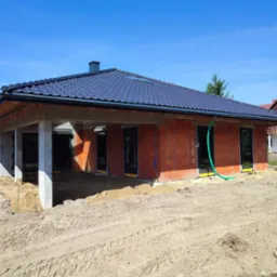 Surowy stan domu jednorodzinnego z czerwonej cegły, z częściowo wykonanym dachem z czarnej dachówki, widoczne okna i drzwi, teren wokół budynku w stanie ziemnym, z śladami opon.