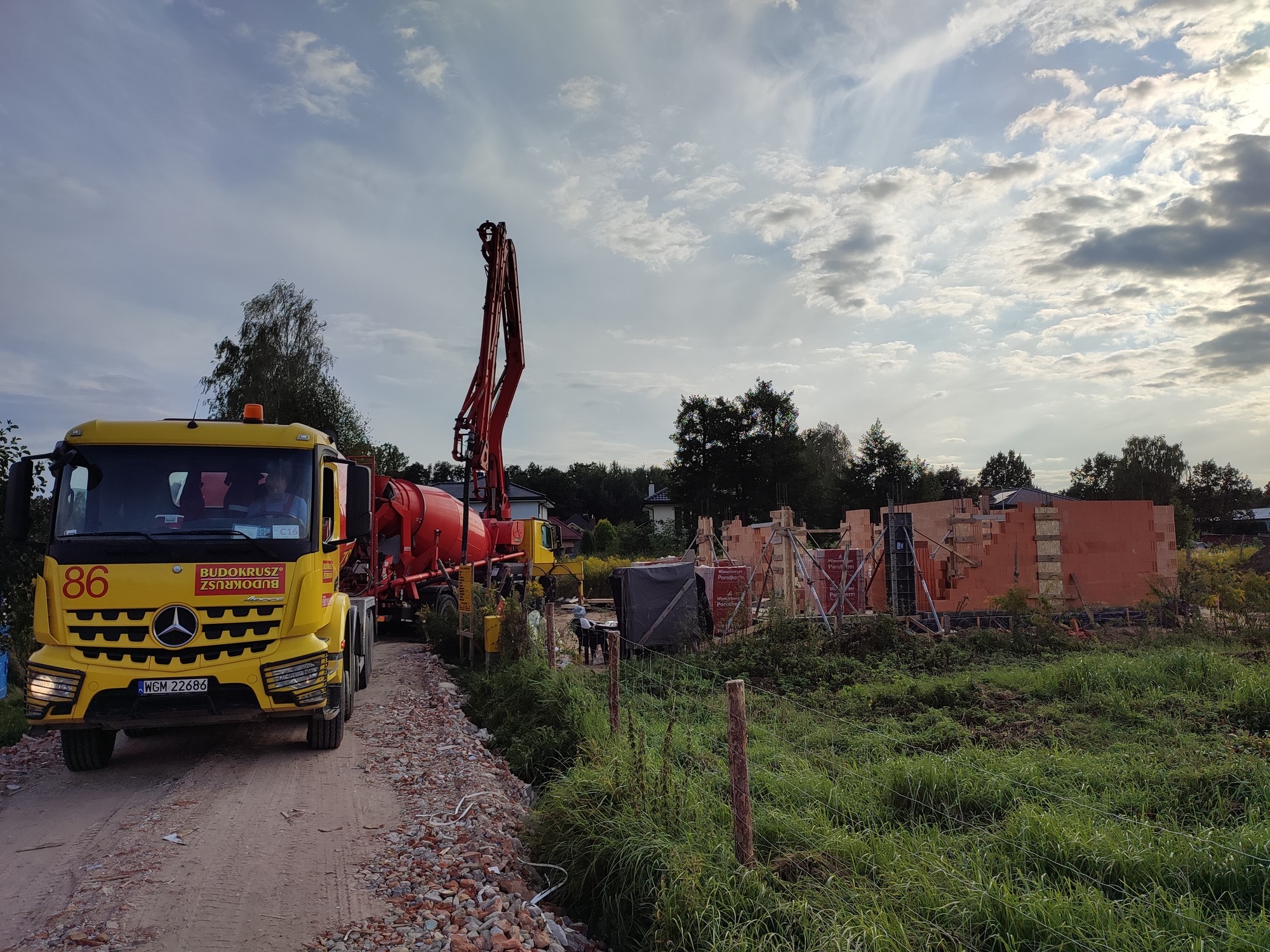 Żółta betoniarka z wysięgnikiem pompuje beton na budowie domu z czerwonej cegły Porotherm, widok z boku na tle zachmurzonego nieba.