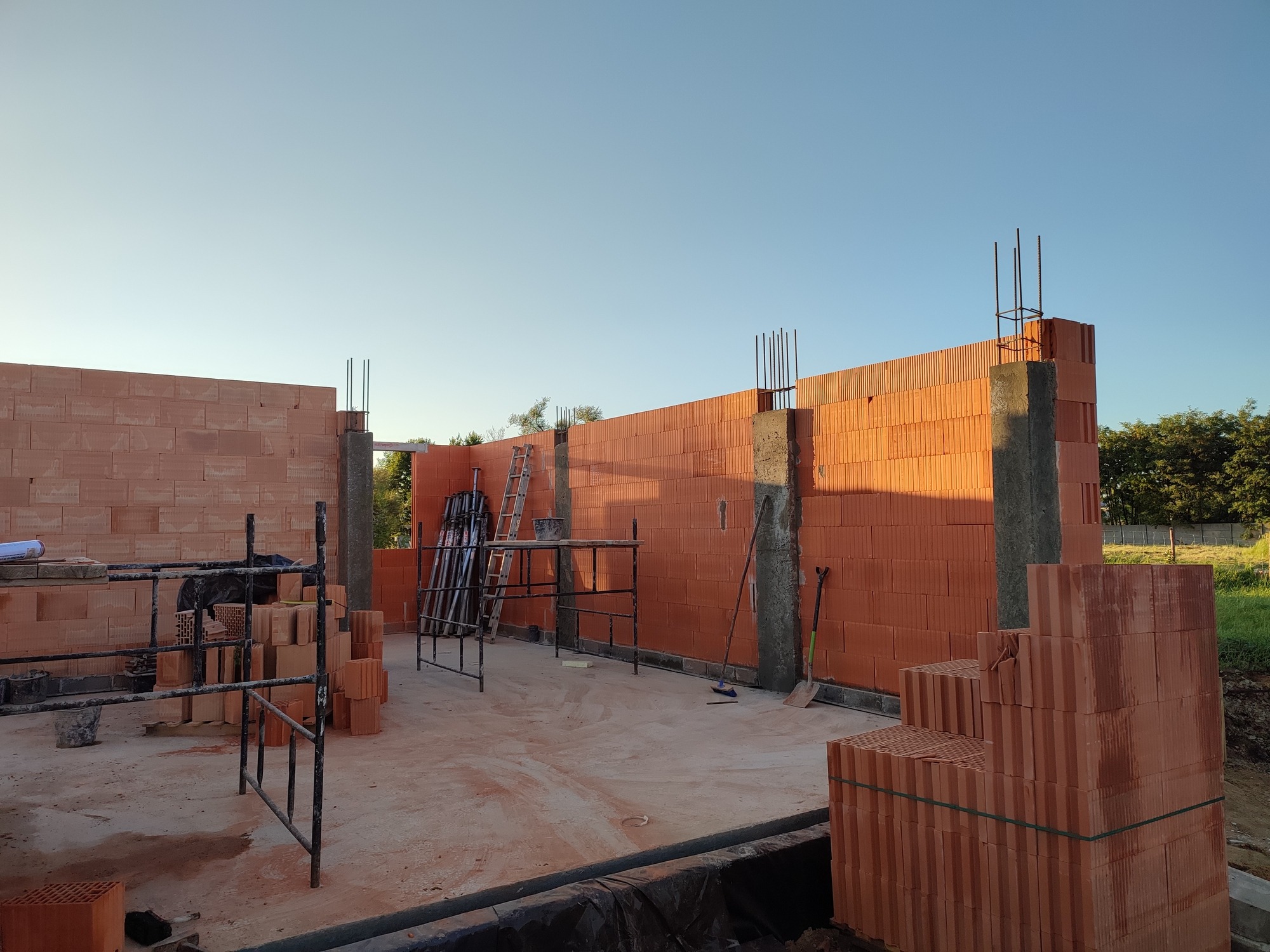 Wznoszenie ścian budynku z czerwonej ceramiki poryzowanej, widoczne szalunki słupów żelbetowych, rusztowania i stosy pustaków na placu budowy, w tle zieleń drzew.