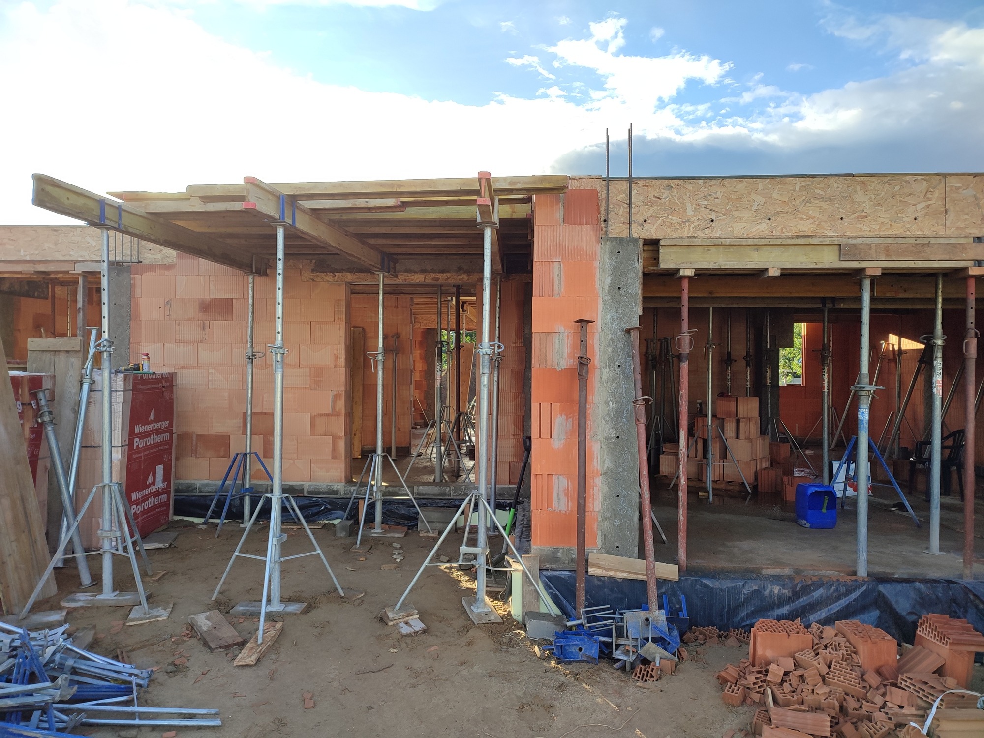 Budowa domu z czerwonej cegły Porotherm w Warszawie, widoczne ściany w trakcie murowania, drewniane szalunki podtrzymywane przez metalowe podpory, na ziemi porozrzucane cegły.