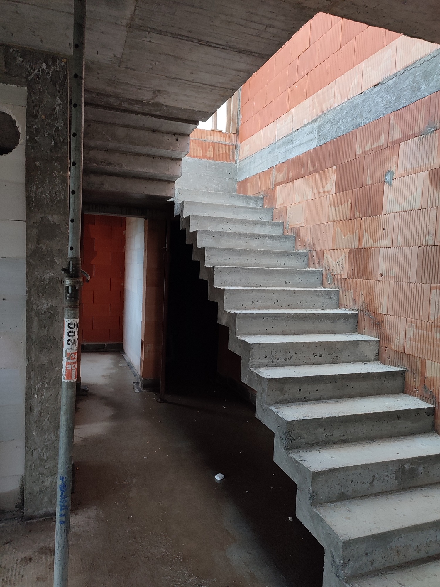 Surowe, betonowe schody wewnętrzne w trakcie budowy, z widocznymi bloczkami z czerwonej cegły i metalowym stemplem budowlanym.