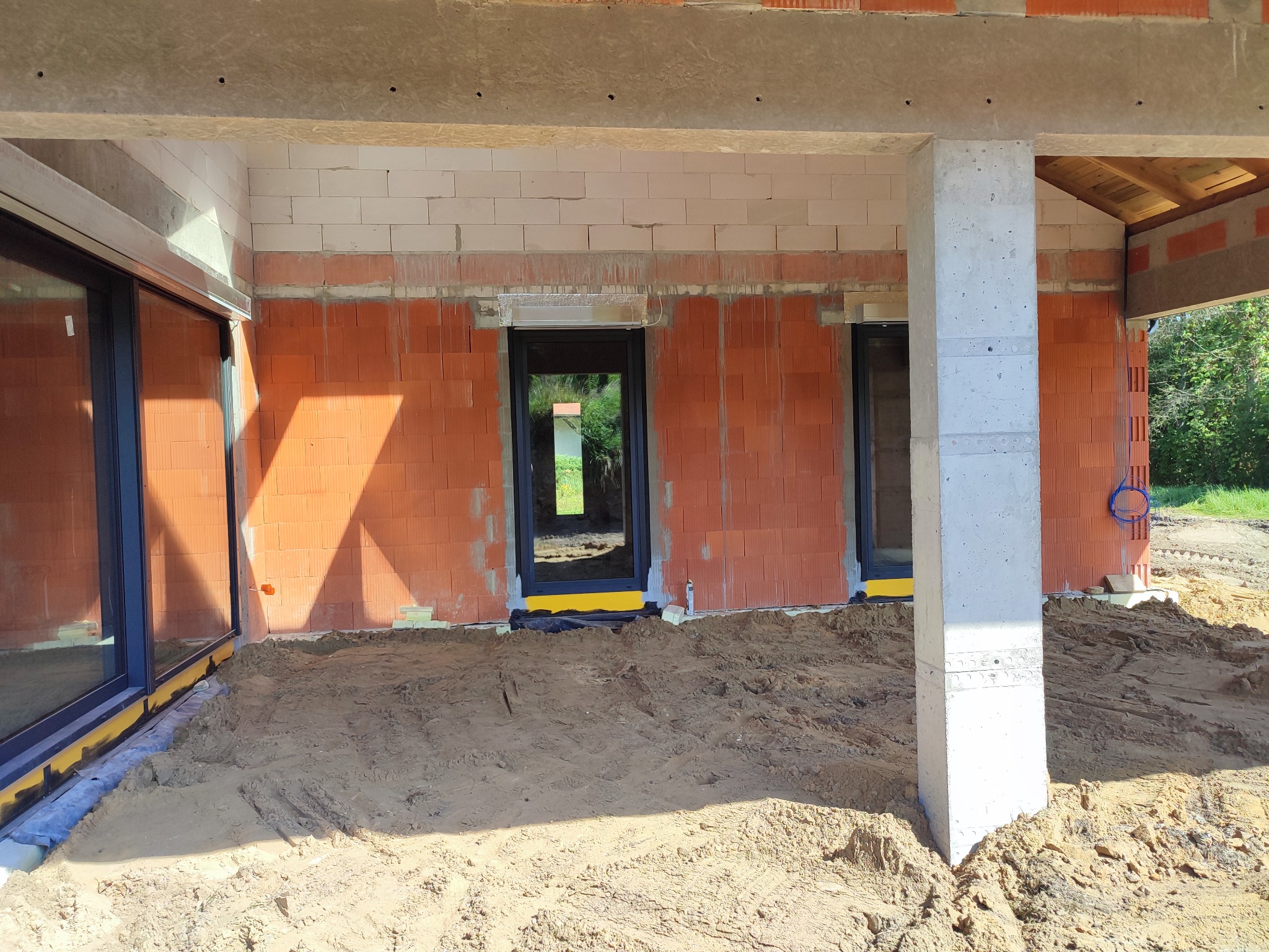 Surowy stan domu murowanego z czerwonej cegły z widocznymi oknami z czarnymi ramami, betonowym słupem i piaskiem na podłożu.