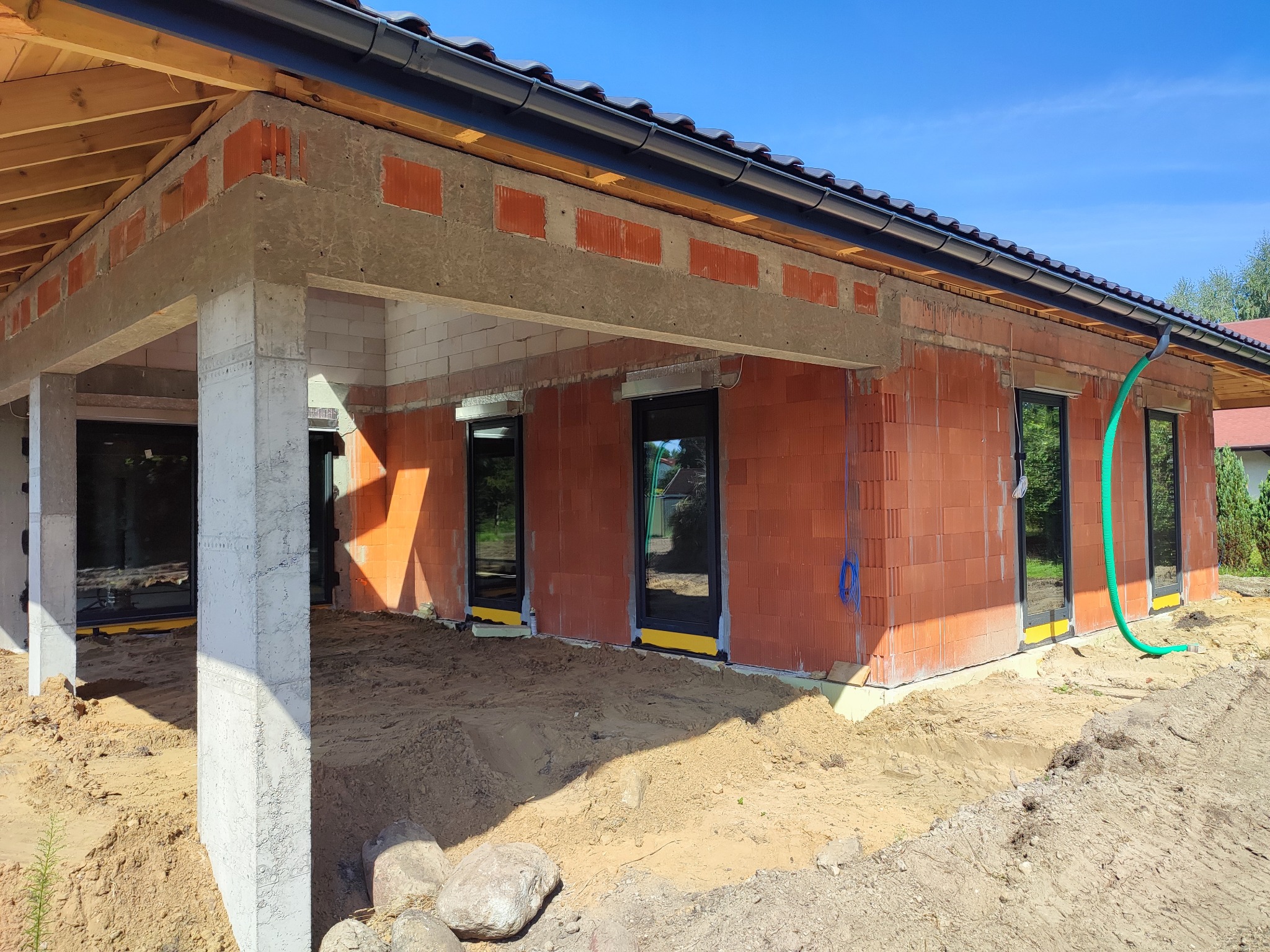 Wznoszenie ścian z czerwonej cegły ceramicznej na budowie domu jednorodzinnego, widoczne okna z czarnymi ramami, betonowe słupy i drewniana konstrukcja dachu pod niebieskim niebem.