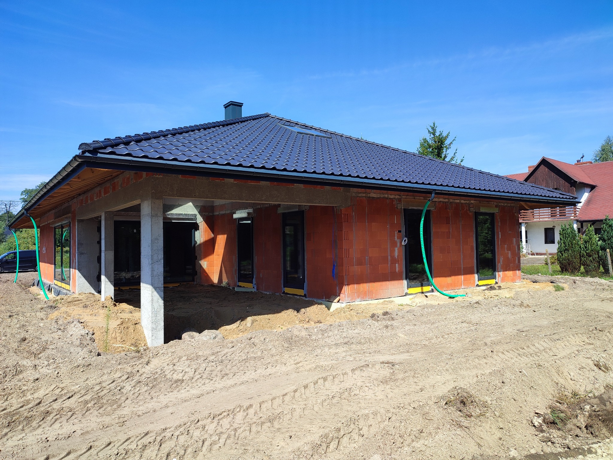 Surowy stan domu jednorodzinnego z czerwonej cegły, z częściowo wykonanym dachem z czarnej dachówki, widoczne okna i drzwi, teren wokół budynku w stanie ziemnym, z śladami opon.