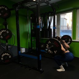 Zuzanna Lipecka - Kobieta wykonuje przysiad ze sztangą w profesjonalnej klatce do ćwiczeń marki Ferros, w tle widoczne obciążenia na stojaku i okno z widokiem na ulicę.