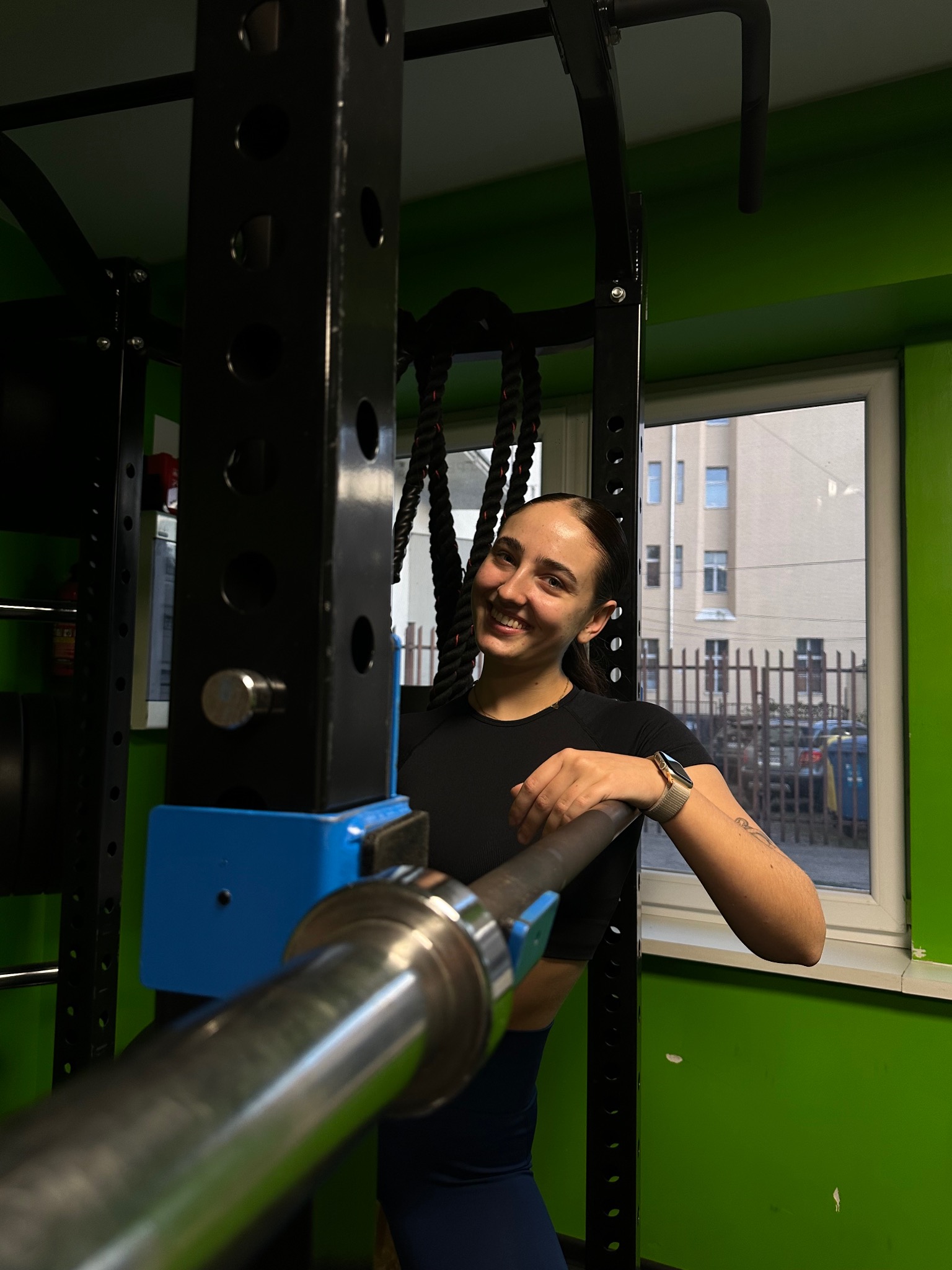 Uśmiechnięta kobieta z zegarkiem i tatuażem na przedramieniu opiera się o sztangę w siłowni z zielonymi ścianami, w tle okno z widokiem na budynek.