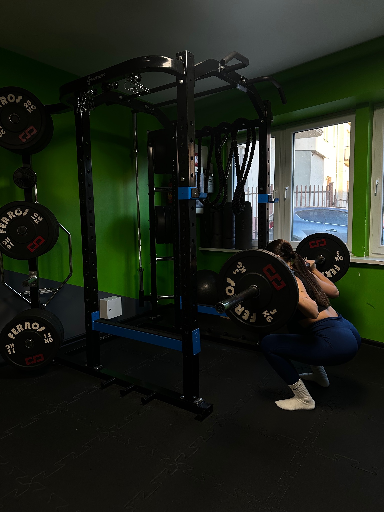 Kobieta wykonuje przysiad ze sztangą w profesjonalnej klatce do ćwiczeń marki Ferros, w tle widoczne obciążenia na stojaku i okno z widokiem na ulicę.