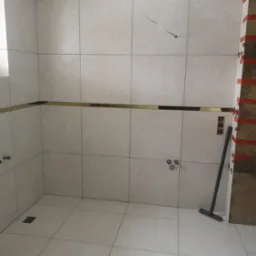 Jasne, kwadratowe płytki podłogowe i ścienne z delikatnym wzorem, układane w łazience, z poziomym, złotym akcentem dekoracyjnym i widocznym odpływem podłogowym oraz narzędziami do układania płytek.