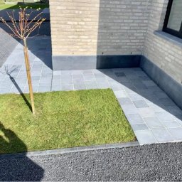 Usługi Budowlane Andrzej Lewandowski - Ułożona szara kostka brukowa tworząca ścieżkę i patio przy budynku z cegły, obok trawnik z młodym drzewkiem, widoczny cień osoby fotografującej.