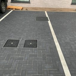 Usługi Budowlane Andrzej Lewandowski - Ułożona ciemnoszara kostka brukowa w jodełkę z białymi liniami wyznaczającymi miejsca parkingowe, widoczne studzienki kanalizacyjne i paleta drewniana przy ścianie budynku.