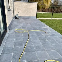 Usługi Budowlane Andrzej Lewandowski - Szare płytki tarasowe ułożone na zewnątrz domu, z żółtym wężem ogrodowym zwiniętym na powierzchni, mur z jasnej cegły w tle, trawnik i płot w oddali.