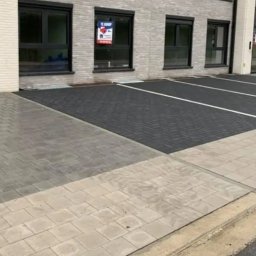 Usługi Budowlane Andrzej Lewandowski - Nowo ułożona kostka brukowa w dwóch odcieniach szarości tworząca parking przed budynkiem z oknami z napisem 'Te Koop'.