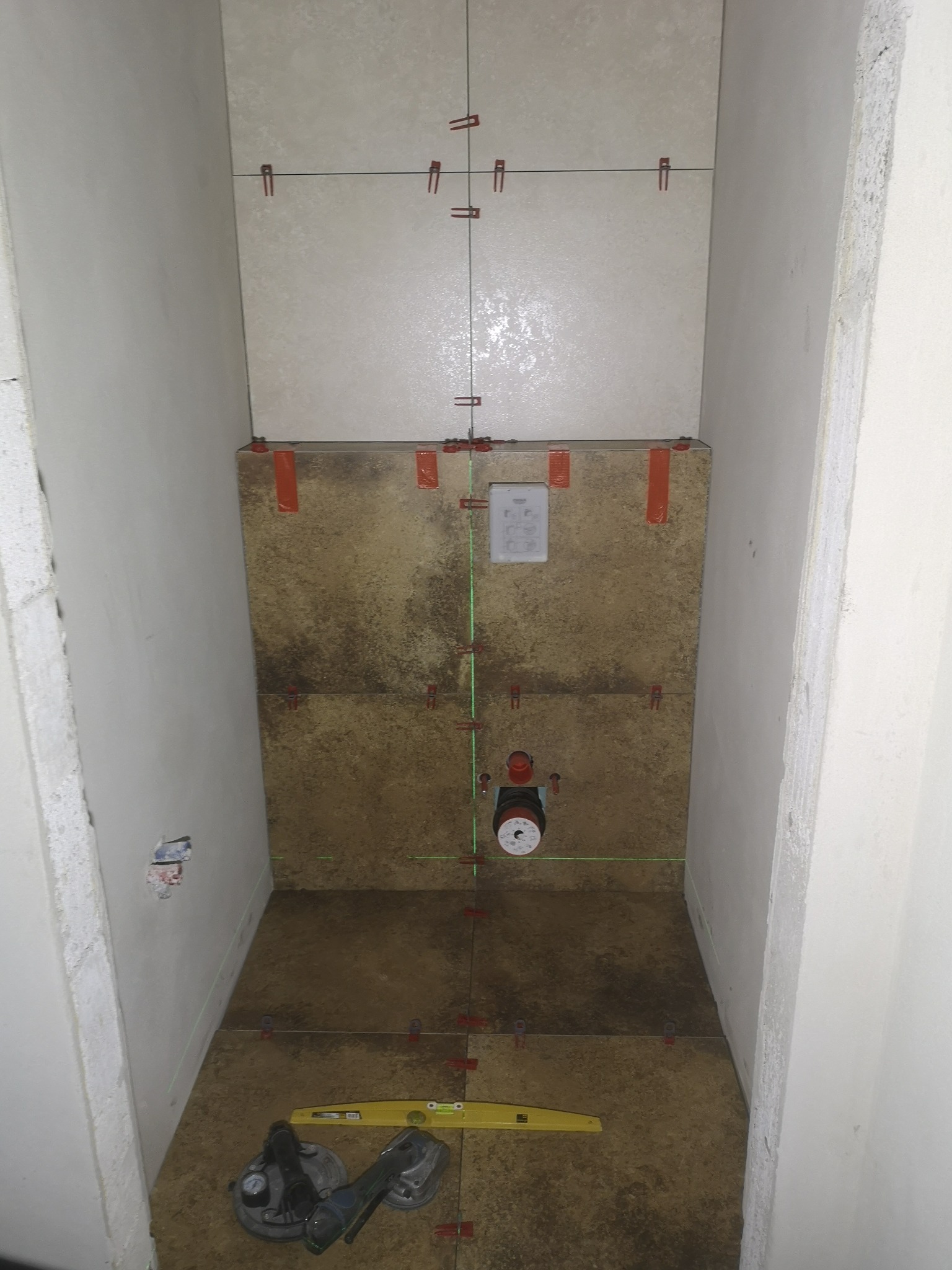 Wnęka w trakcie wykańczania płytkami w odcieniach beżu i brązu, widoczne poziomice laserowe, przyssawka do płytek i krzyżyki dystansowe, przygotowanie do montażu armatury.
