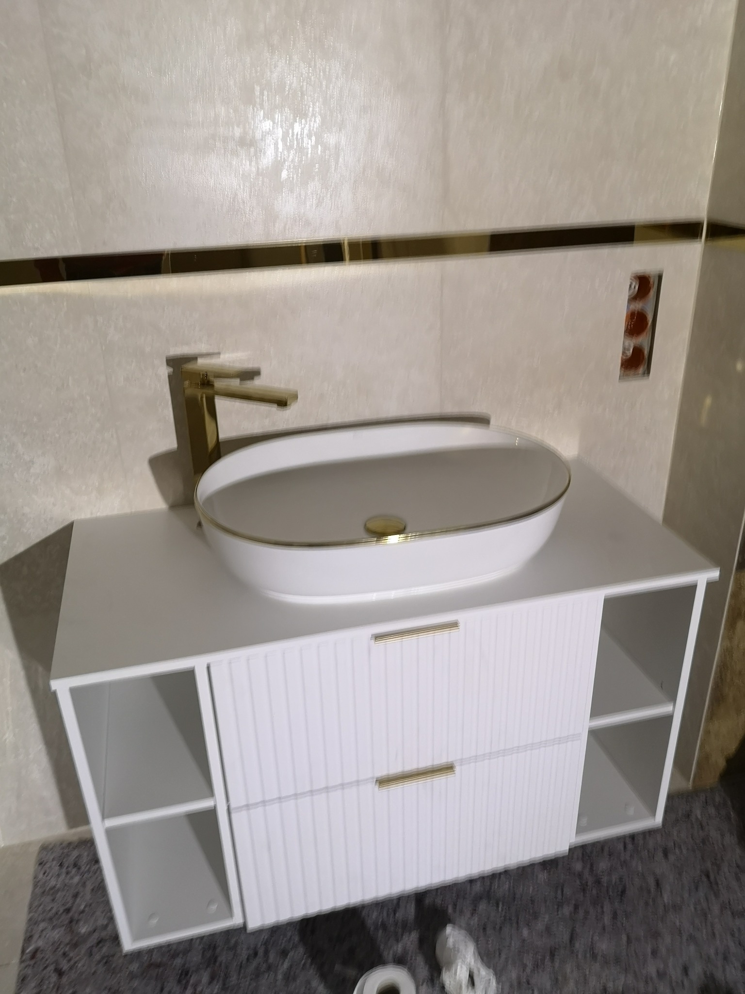 Nowoczesna łazienka: biała szafka z umywalką nablatową o owalnym kształcie z delikatnym złotym akcentem, złota bateria, kremowe płytki z poziomym paskiem dekoracyjnym w kolorze złota, widoczne...