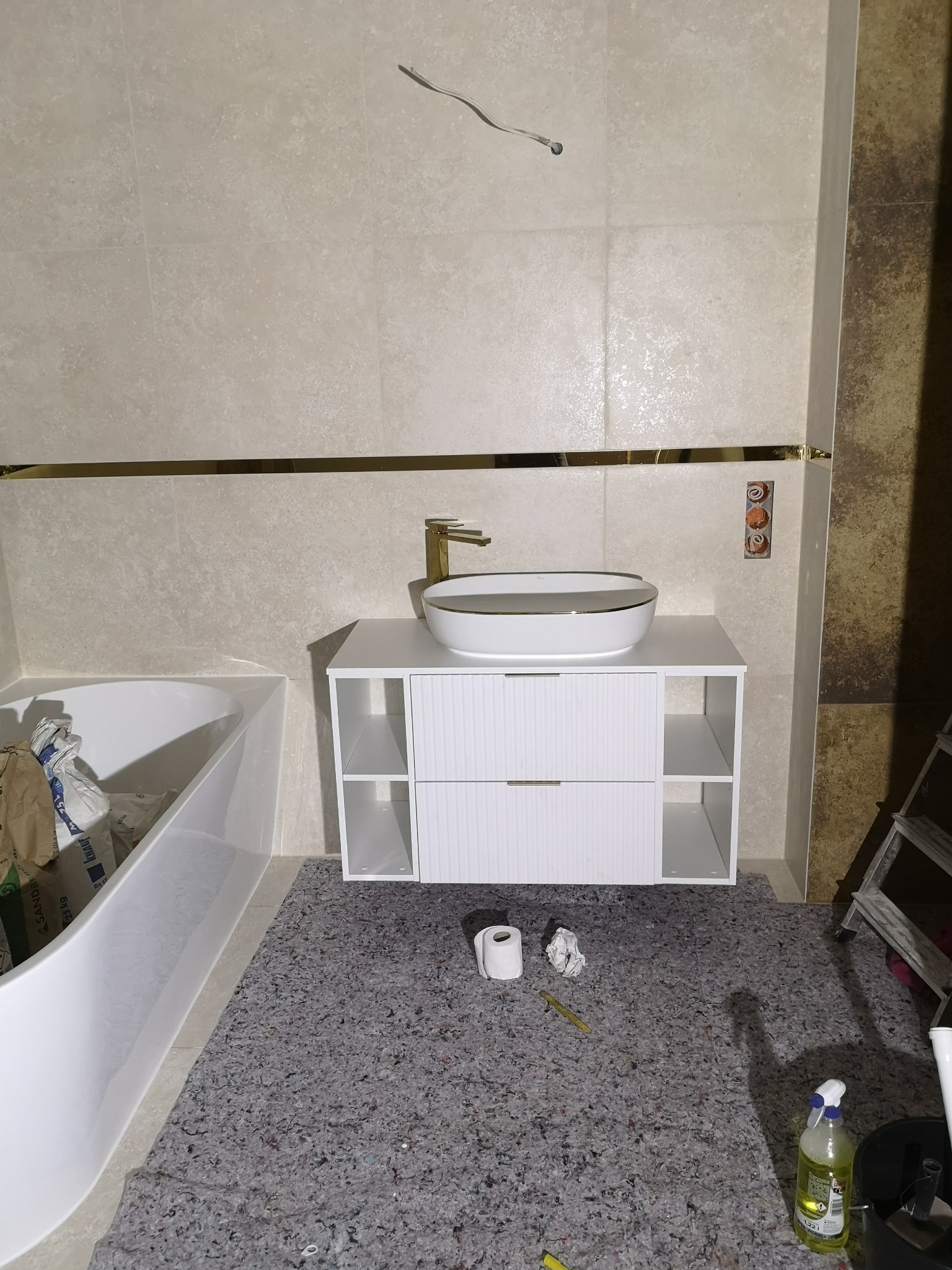 Biała umywalka nablatowa z złotą armaturą na szafce z frezowanymi frontami w łazience z beżowymi płytkami i złotym akcentem, widoczne worki z zaprawą klejową przy wannie i zabezpieczona podłoga.