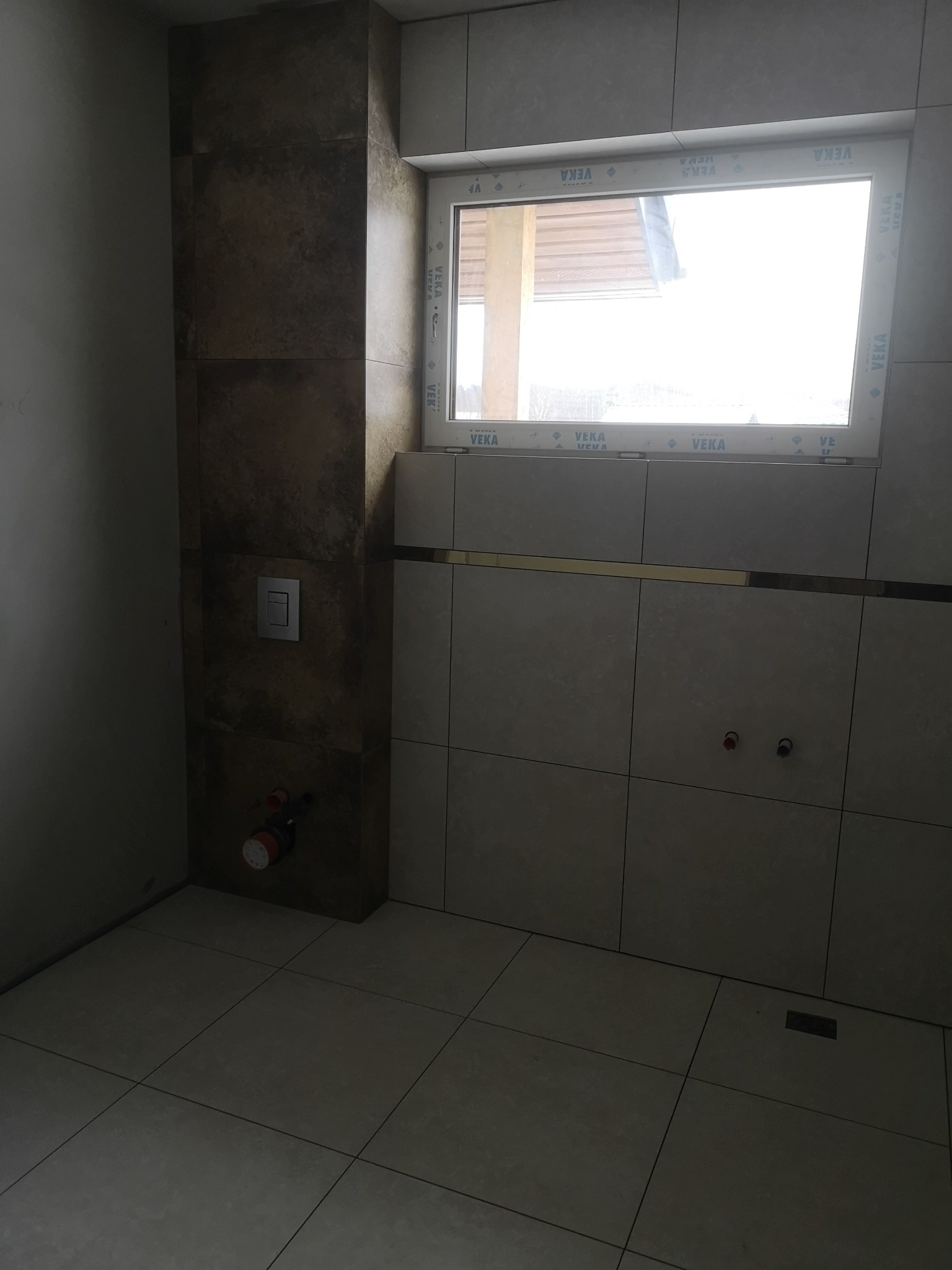 Wykończona łazienka w Słupsku, z jasnymi płytkami na ścianach i podłodze, okno PCV marki VEKA, widoczny fragment instalacji wodnej i spłuczka podtynkowa na ścianie z brązowymi płytkami.