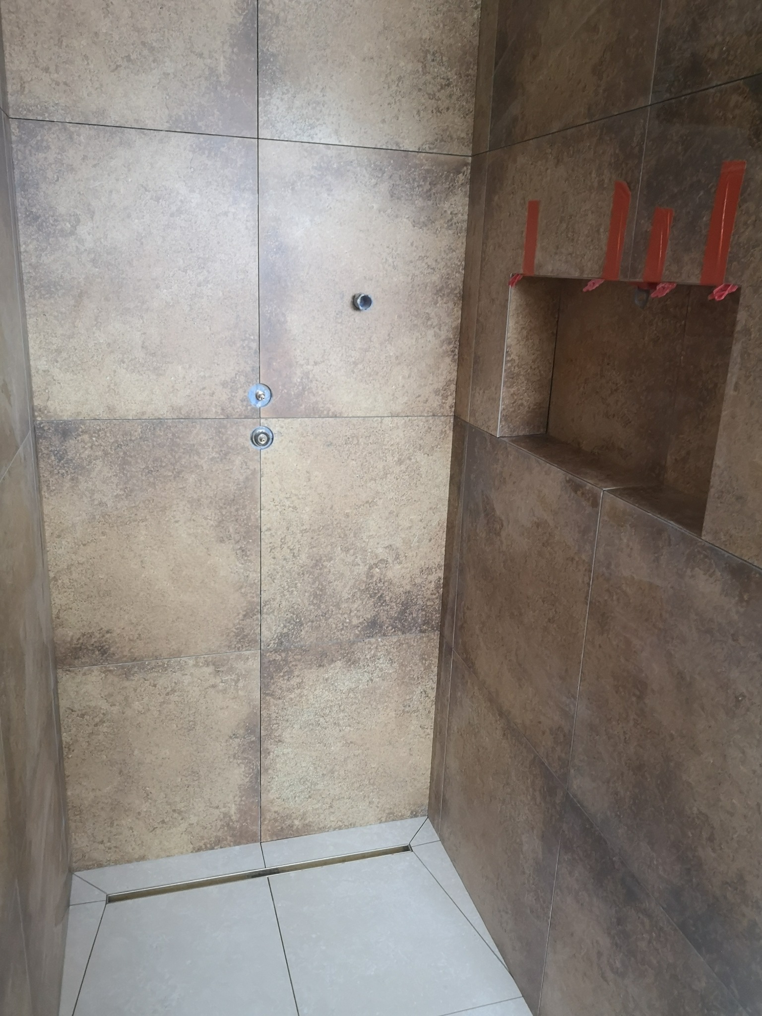 Ściana wyłożona brązowo-beżowymi płytkami imitującymi kamień, z wnęką ozdobioną pomarańczowymi klinami dystansowymi i dwoma otworami na armaturę, widoczny odpływ liniowy w podłodze.