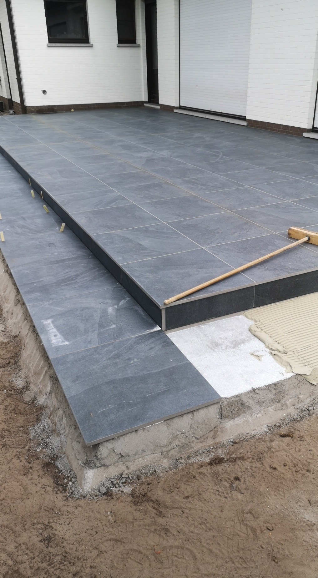 Szara, płytkowa nawierzchnia tarasu w trakcie budowy, z widocznym fragmentem podłoża i warstwą kleju, oraz szczotką opartą o krawędź.