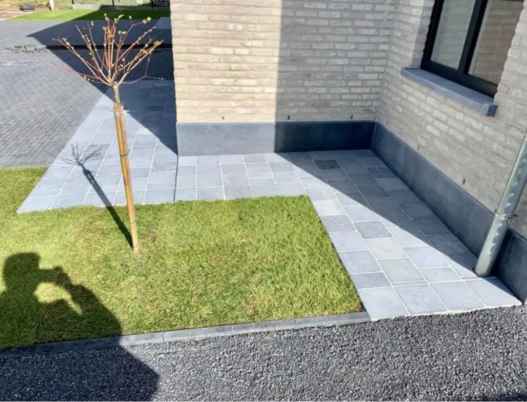 Ułożona szara kostka brukowa tworząca ścieżkę i patio przy budynku z cegły, obok trawnik z młodym drzewkiem, widoczny cień osoby fotografującej.