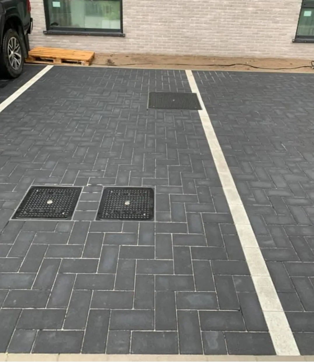 Ułożona ciemnoszara kostka brukowa w jodełkę z białymi liniami wyznaczającymi miejsca parkingowe, widoczne studzienki kanalizacyjne i paleta drewniana przy ścianie budynku.