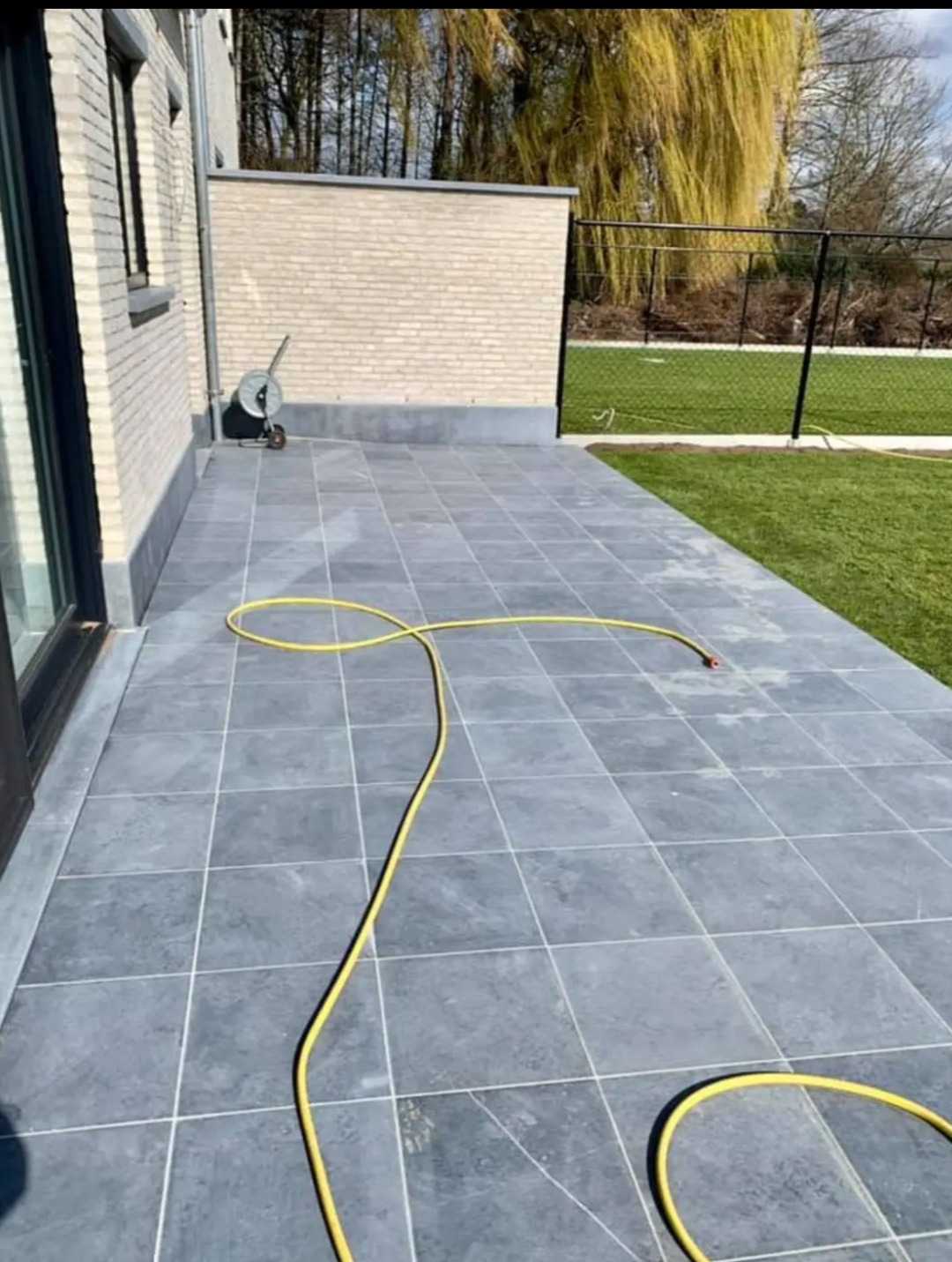 Szare płytki tarasowe ułożone na zewnątrz domu, z żółtym wężem ogrodowym zwiniętym na powierzchni, mur z jasnej cegły w tle, trawnik i płot w oddali.
