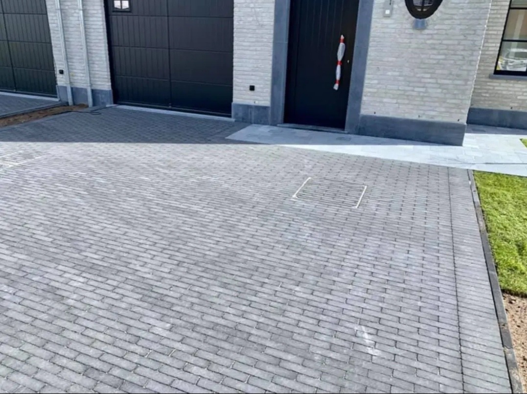 Nowo ułożona, szara kostka brukowa na podjeździe przed domem z dwoma garażami i czarnymi drzwiami wejściowymi, widoczna kratka kanalizacyjna wkomponowana w nawierzchnię.