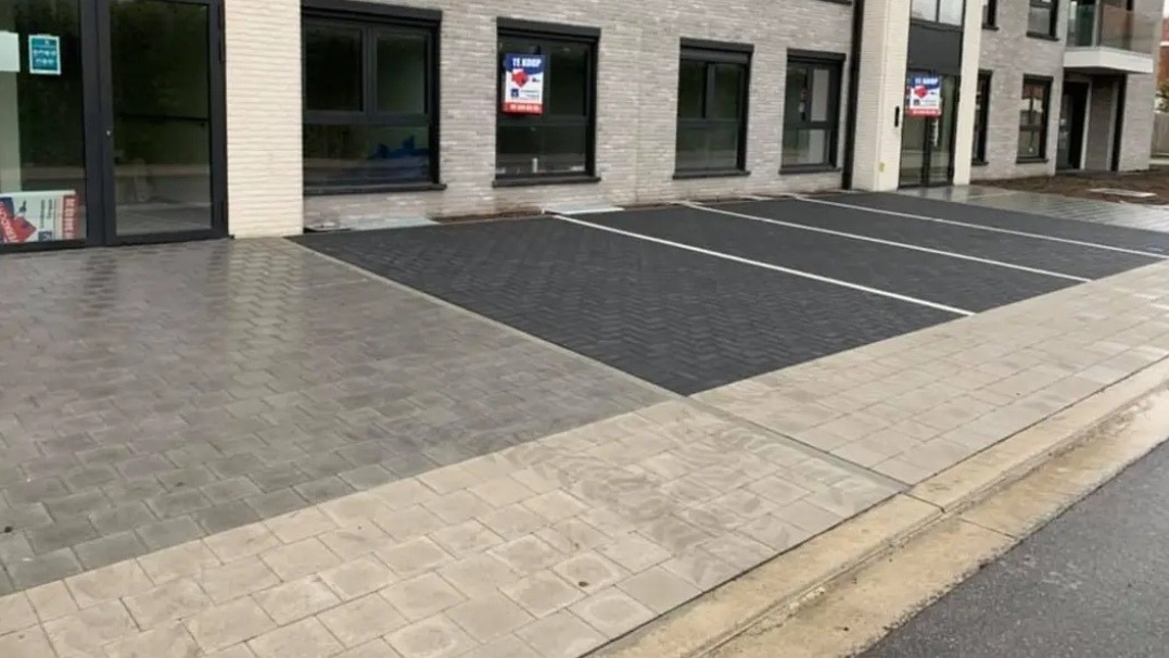 Nowo ułożona kostka brukowa w dwóch odcieniach szarości tworząca parking przed budynkiem z oknami z napisem 'Te Koop'.