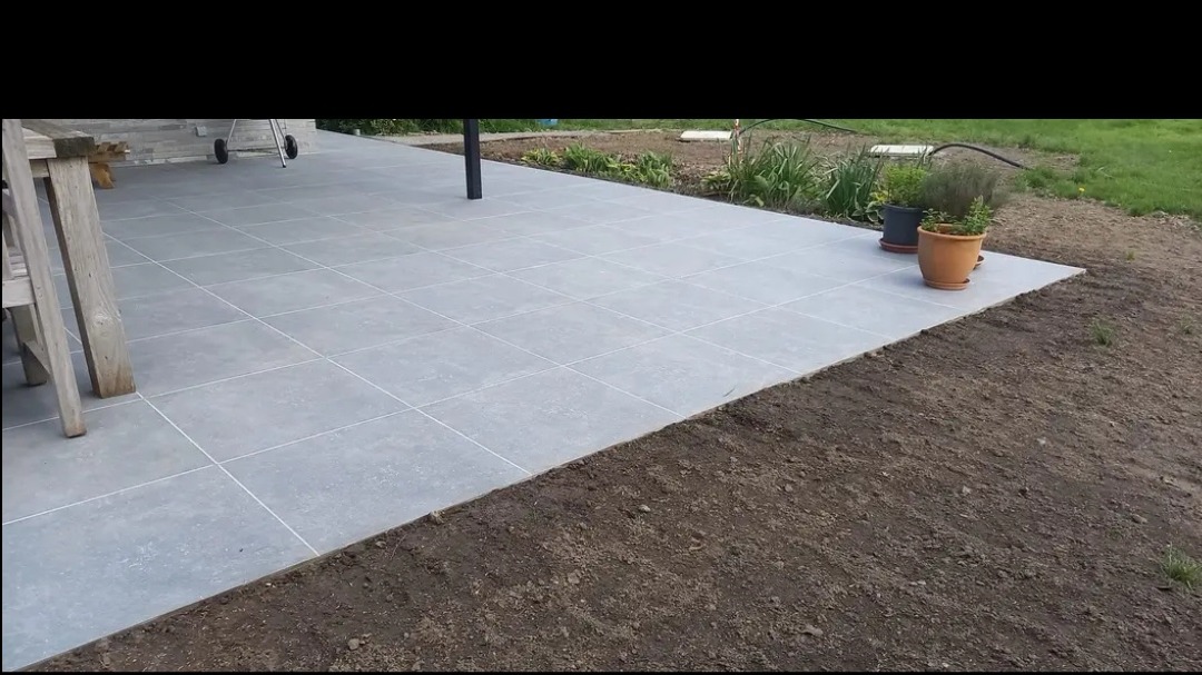 Szara, kwadratowa nawierzchnia z płytek ułożona na zewnątrz, przylegająca do świeżo przekopanej ziemi, z doniczkami i grillem w tle.