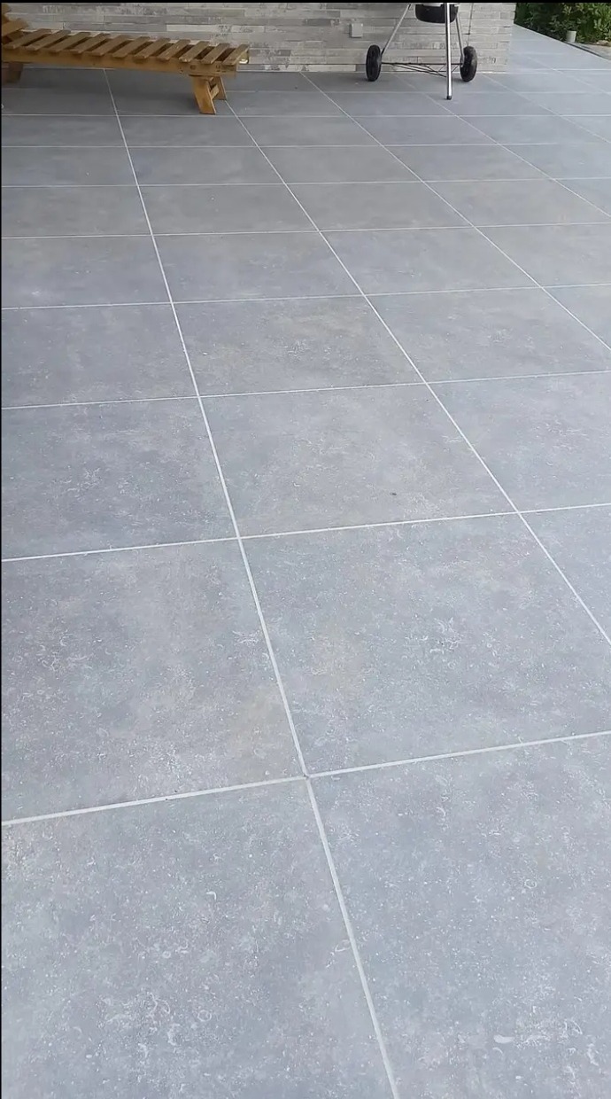 Świeżo ułożona, jasnoszara kostka brukowa na tarasie, z widocznym drewnianym leżakiem i grillem w tle.