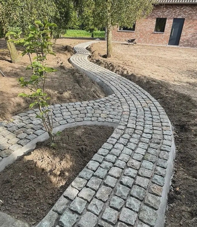 Kręta ścieżka z kostki brukowej prowadząca przez ogród, z młodym drzewkiem po lewej stronie i domem z cegły w tle.