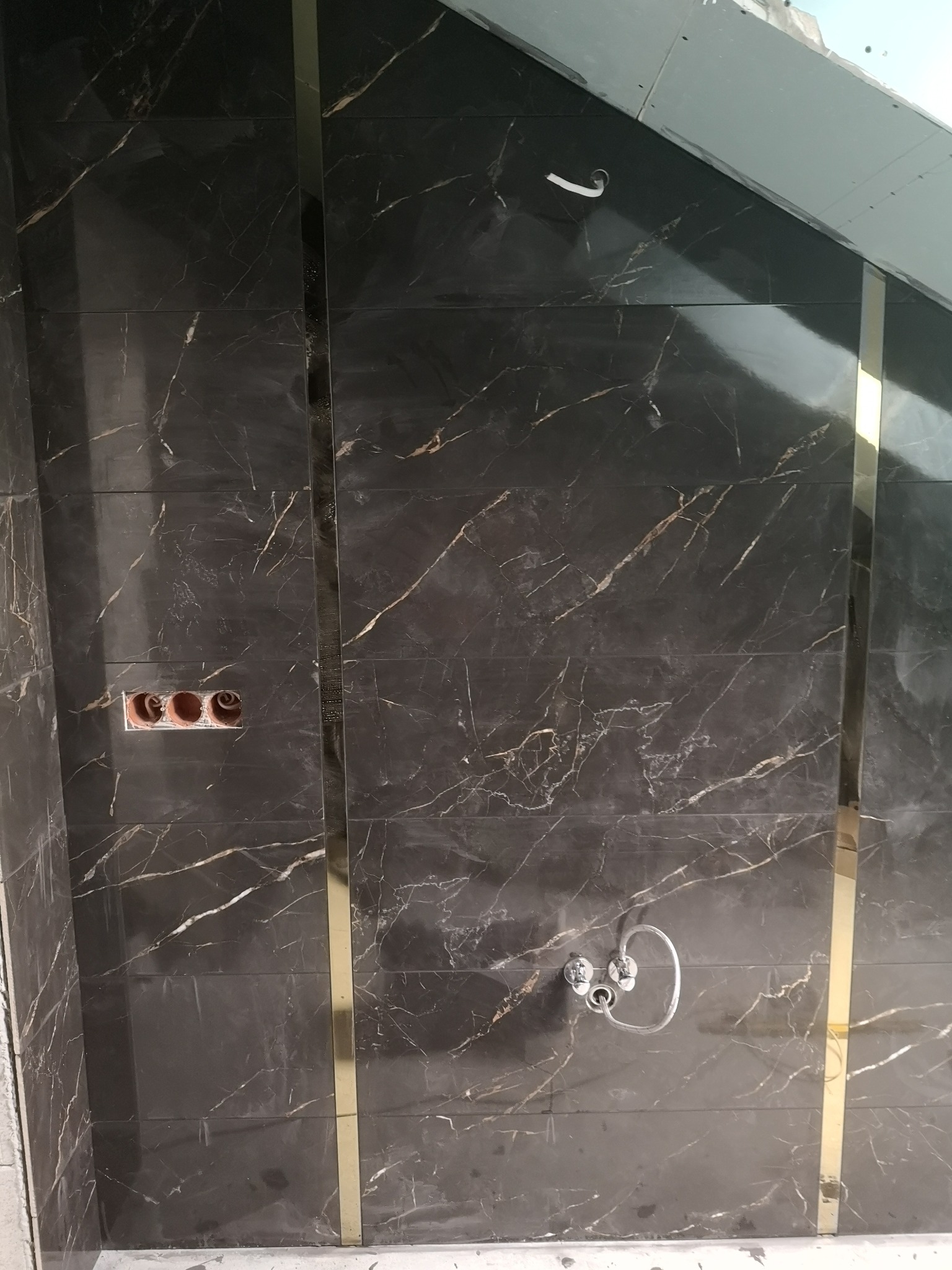 Ściana łazienki wyłożona czarnymi płytkami imitującymi marmur, z ozdobnymi złotymi listwami i instalacją wodno-kanalizacyjną w trakcie montażu.