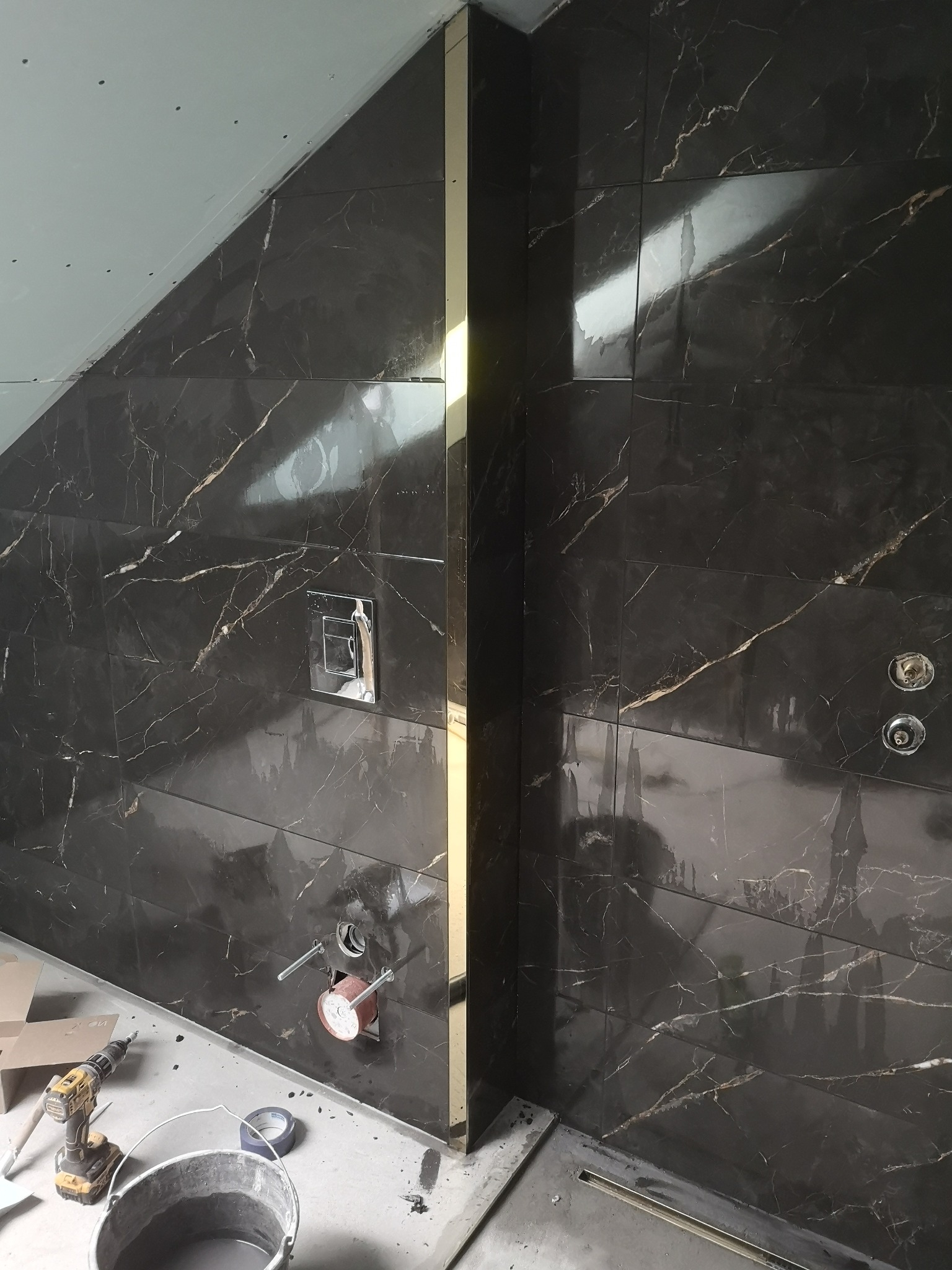 Częściowo wykończona łazienka z ciemnymi, marmurkowymi płytkami, złotą listwą narożną, widocznymi elementami instalacji wodnej i narzędziami glazurniczymi na podłodze.