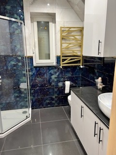 Nowoczesna łazienka z kabiną prysznicową w rogu, ciemnymi płytkami imitującymi marmur z niebieskimi żyłami na ścianach, białymi szafkami pod umywalką i grzejnikiem drabinkowym w kolorze złotym.