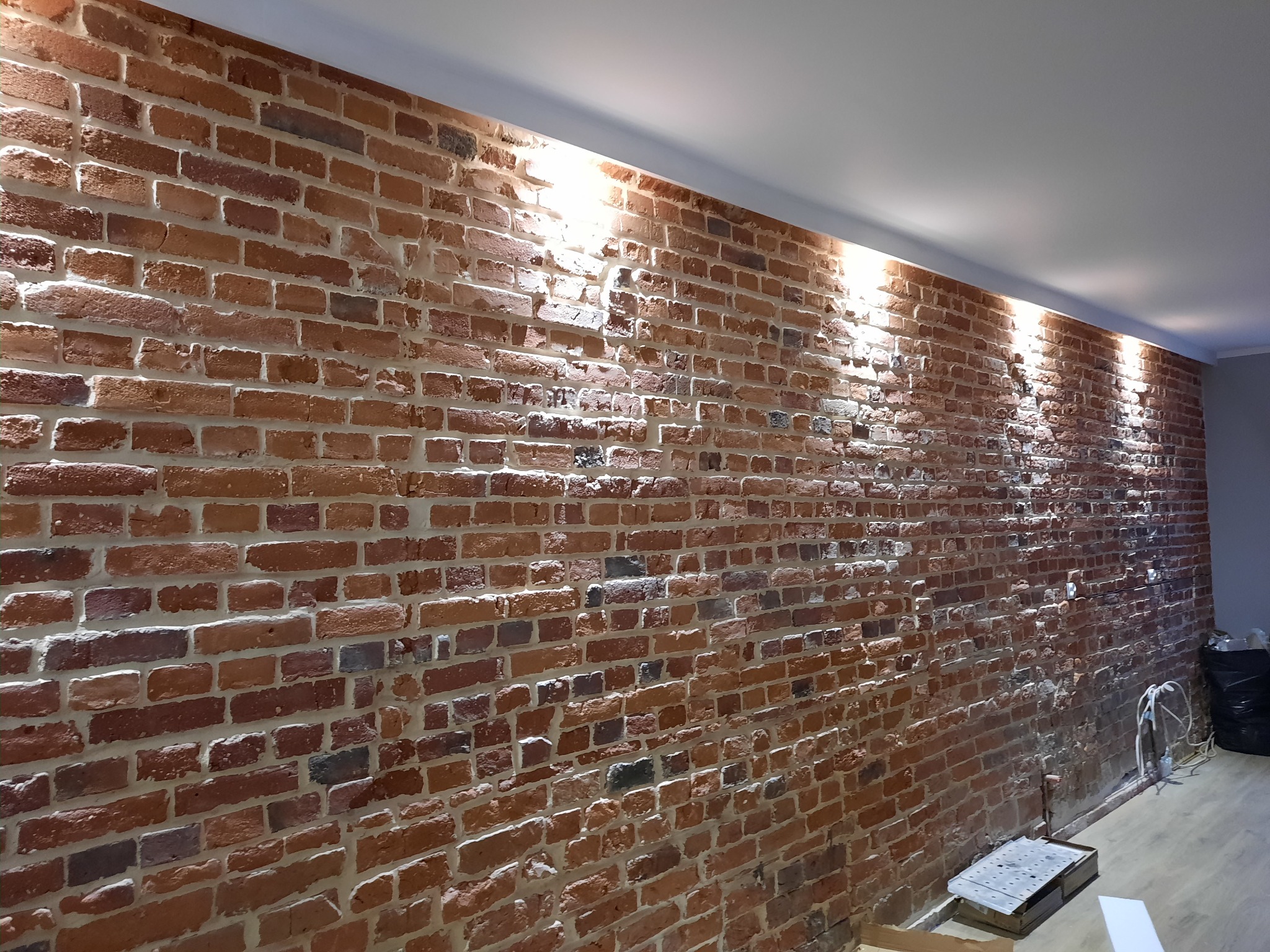 Ściana z czerwonej cegły z widocznymi fugami, oświetlona od góry światłem LED, z elementami instalacji elektrycznej i narzędziami w dolnej części kadru.