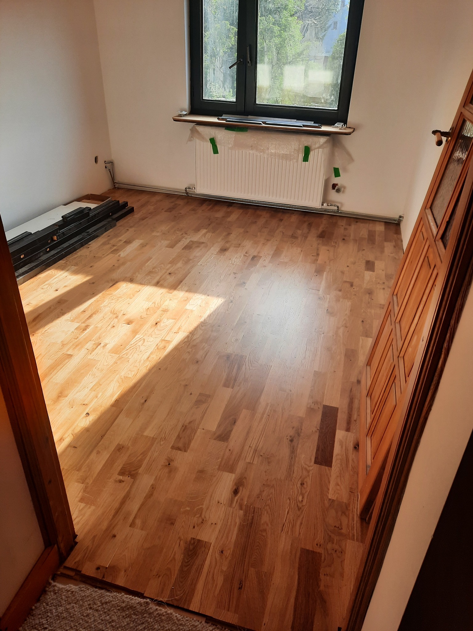 Wykończona podłoga z paneli w jasnym odcieniu drewna w pokoju z oknem i grzejnikiem zabezpieczonym folią, panele ułożone równolegle do okna, przy ścianie leżą paczki paneli gotowe do użycia.