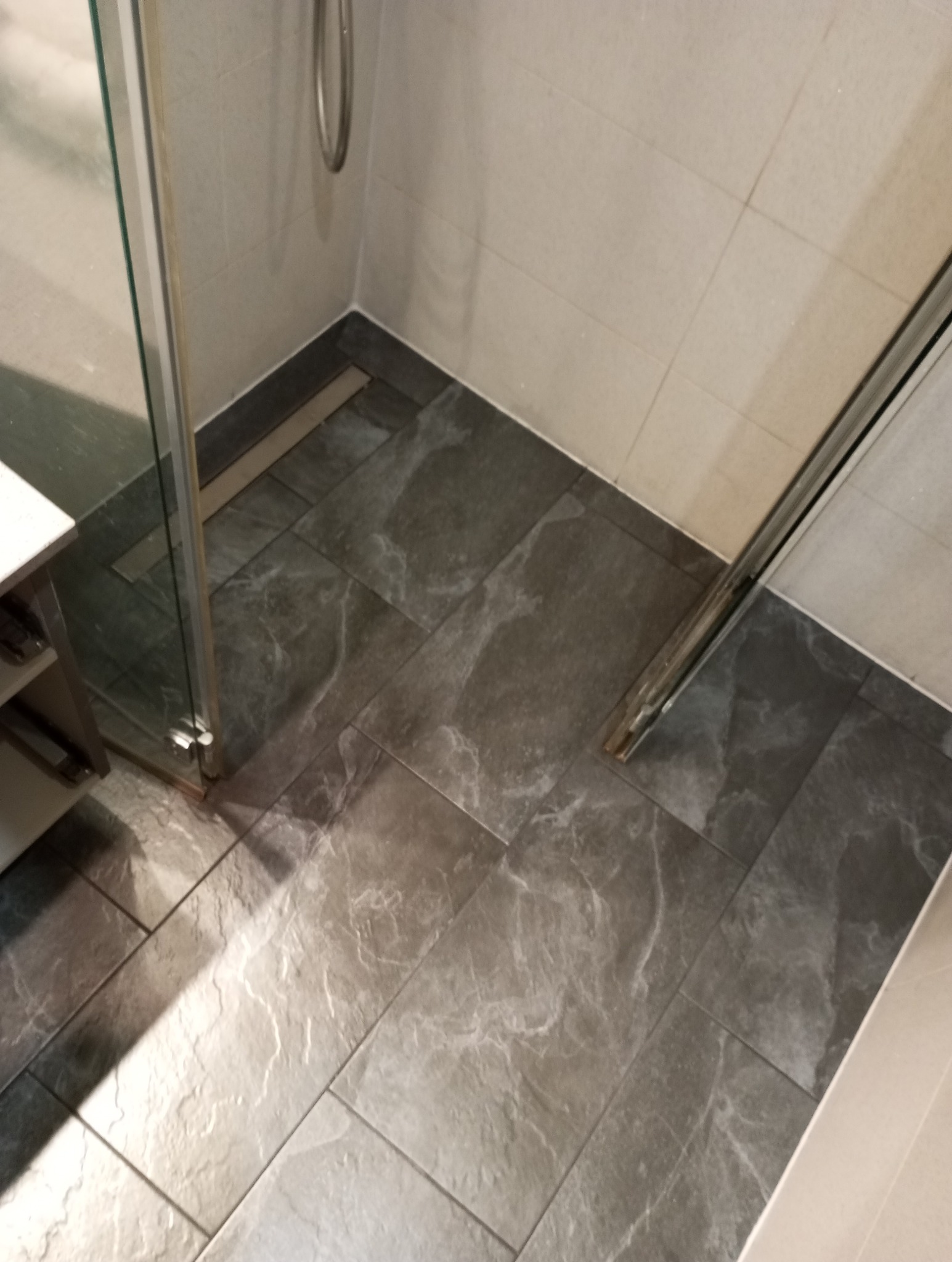 Wykończona strefa prysznicowa z szarymi płytkami imitującymi marmur, kabina prysznicowa z przezroczystego szkła, odpływ liniowy przy ścianie.