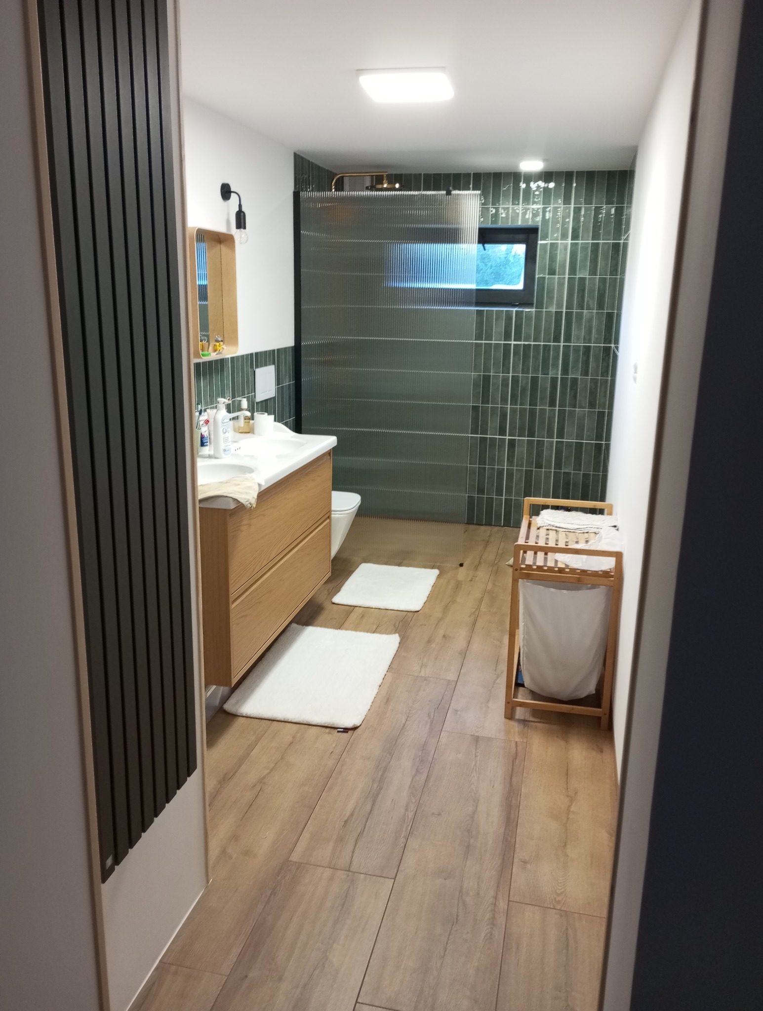 Nowoczesna łazienka z drewnianą szafką pod umywalką, zielonymi płytkami na ścianach i kabiną prysznicową z ryflowanego szkła. Widoczne białe dywaniki i drewniana podłoga.