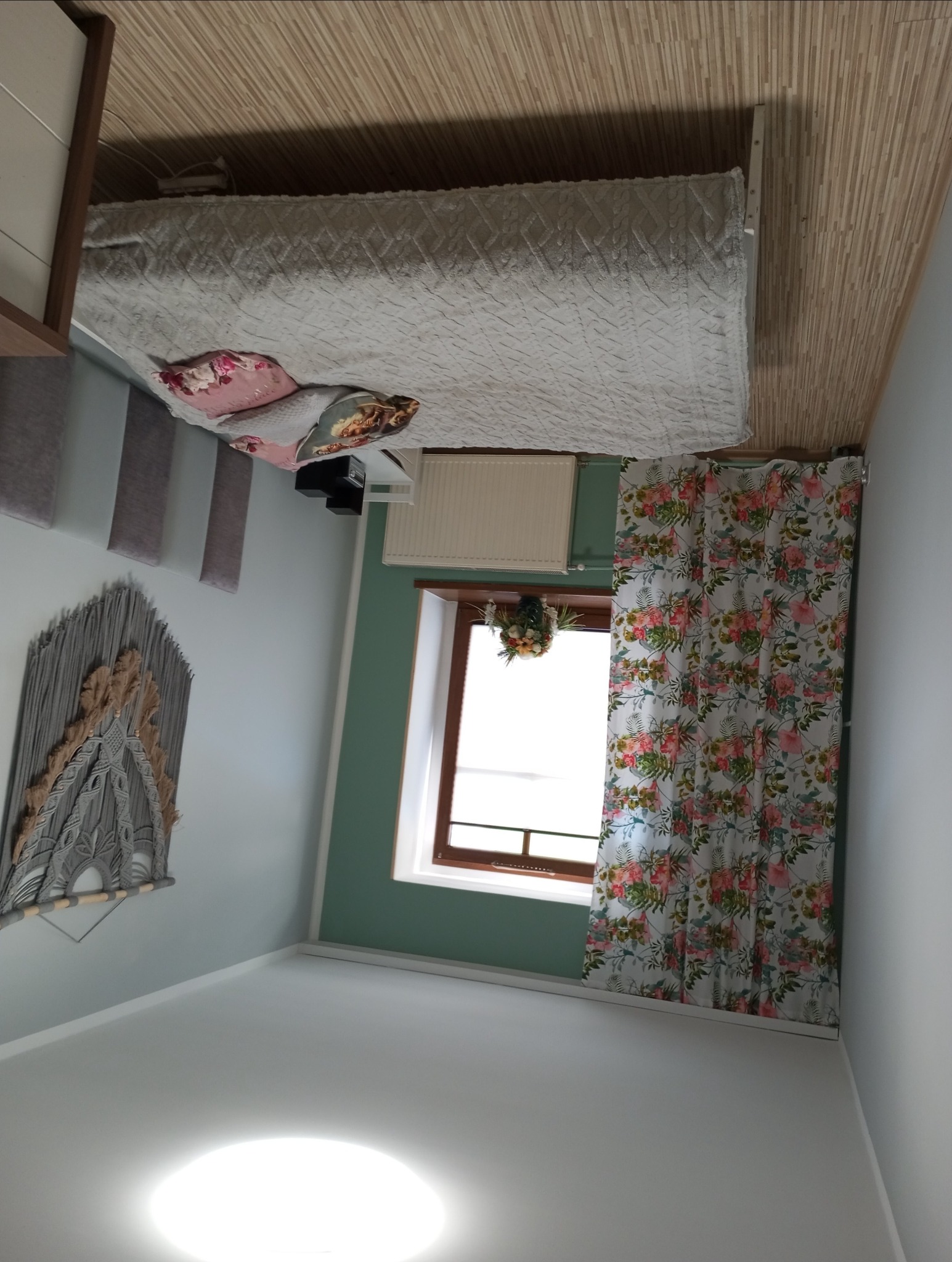 Jasne wnętrze sypialni po remoncie: białe ściany, drewniana podłoga, okno z kwiatowymi zasłonami, dekoracyjny makramowy panel na ścianie i łóżko z wzorzystą narzutą.