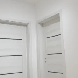 Dwa białe, nowoczesne drzwi wewnętrzne z poziomymi, ciemnymi wstawkami, zamontowane w białej ścianie, widoczne otwory na klamki.