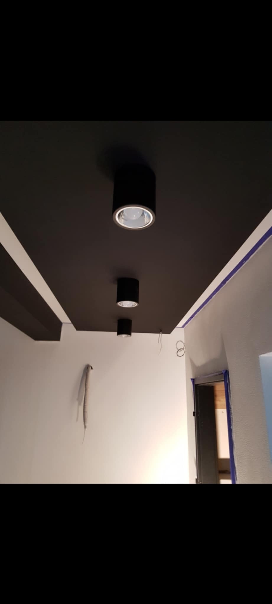 Czarny sufit z trzema okrągłymi, wpuszczanymi lampami, białe ściany z niebieską taśmą malarską przy suficie, widoczne przewody i otwór drzwiowy.