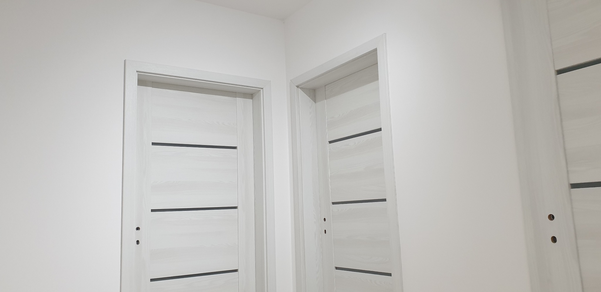 Dwa białe, nowoczesne drzwi wewnętrzne z poziomymi, ciemnymi wstawkami, zamontowane w białej ścianie, widoczne otwory na klamki.