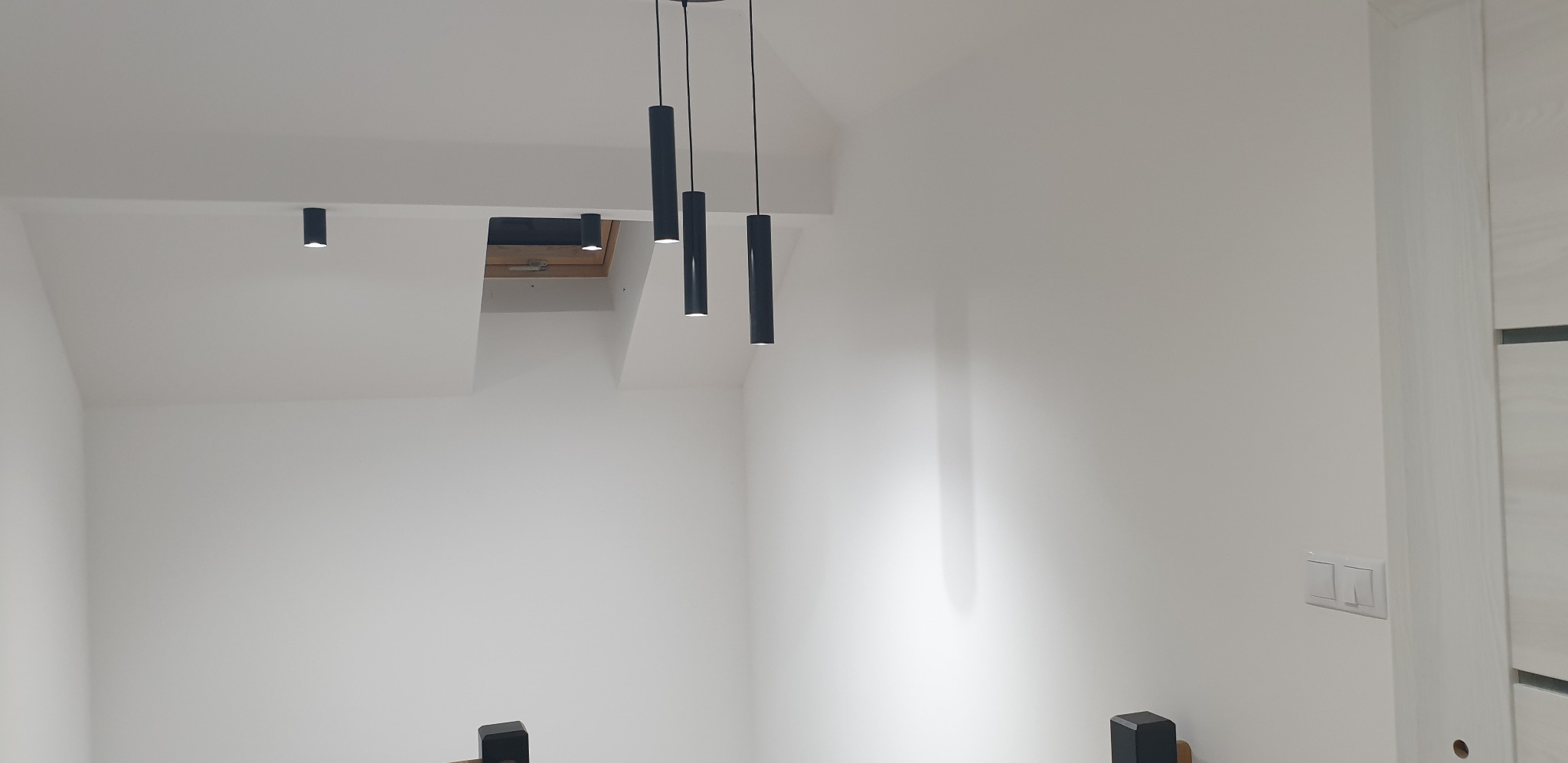 Wnętrze poddasza ze skośnym sufitem, pomalowane na biało, z trzema wiszącymi czarnymi lampami cylindrycznymi i lampą sufitową w pobliżu okna dachowego.