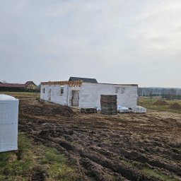 Firma Budowlana Jarosław Spiczak Brzeziński - Wznoszenie ścian parterowego domu z białych bloczków, widoczne palety i paczki materiałów budowlanych na błotnistym terenie działki.