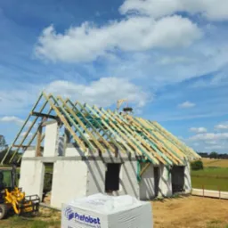 Wznoszenie konstrukcji dachu na surowym budynku z bloczków betonowych, widoczny miniładowarka i paleta bloczków Prefabet