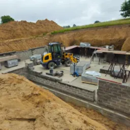 Budowa fundamentów domu z bloczków betonowych, widoczny żółty miniładowarka, wykop pod fundamenty i przygotowane podpory pod szalunek.