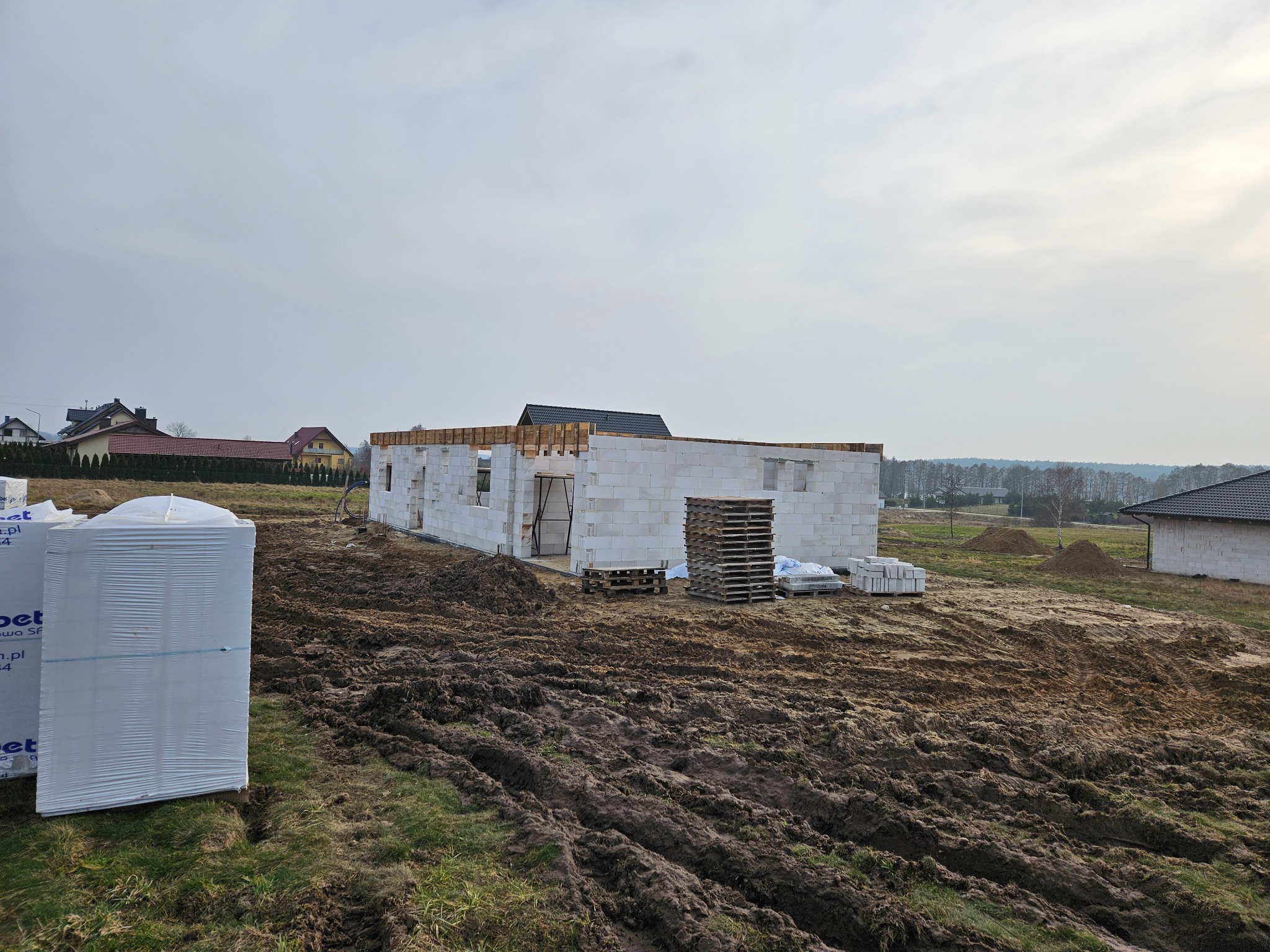 Wznoszenie ścian parterowego domu z białych bloczków, widoczne palety i paczki materiałów budowlanych na błotnistym terenie działki.