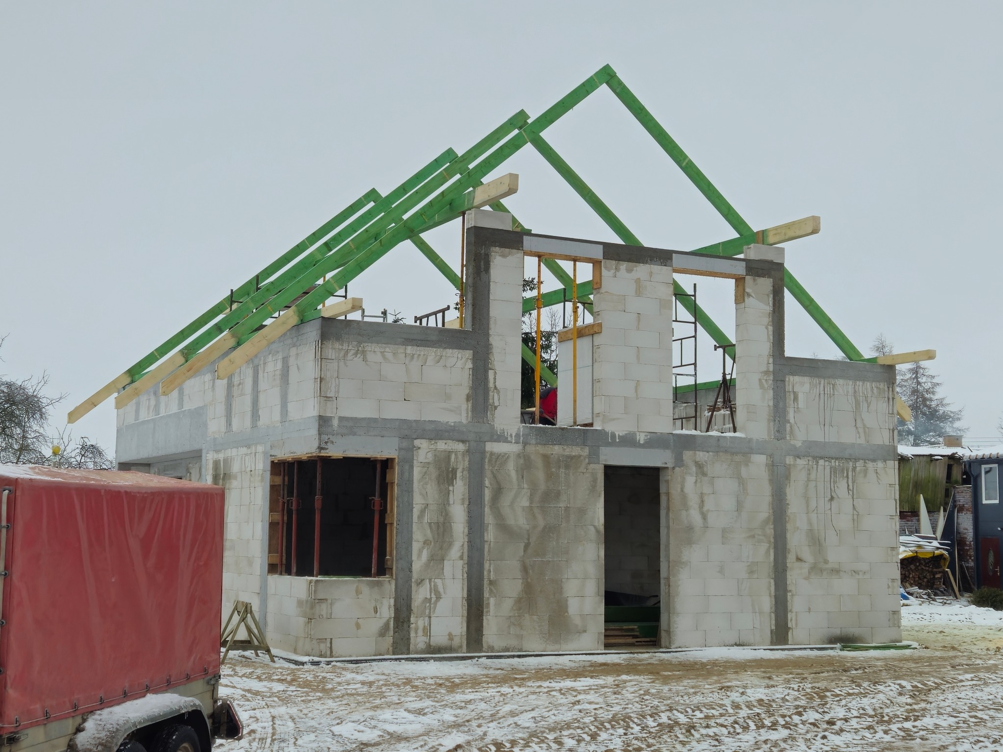 Surowy stan domu z betonu komórkowego w Bytowie, z zielonymi elementami konstrukcji dachu i czerwonym przyczepą z plandeką w zimowej scenerii.