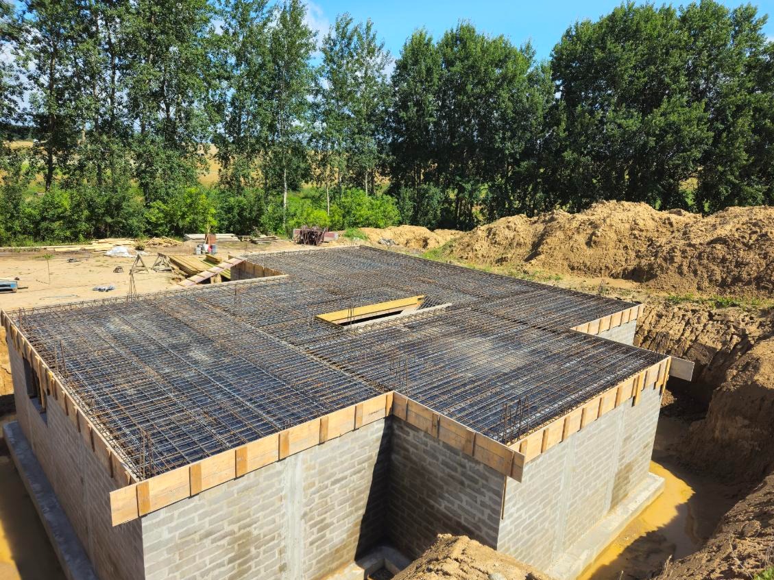 Wylany fundament zbrojony stalą, widoczne ściany fundamentowe z bloczków betonowych, wykop pod fundament, w tle zieleń drzew.