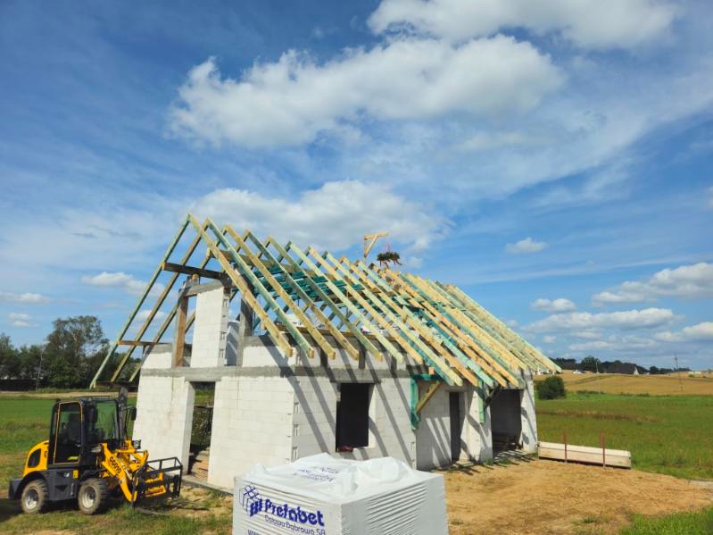 Wznoszenie konstrukcji dachu na surowym budynku z bloczków betonowych, widoczny miniładowarka i paleta bloczków Prefabet